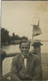 A young Aldo Leopold poses smiling wearing a jacket and bow tie while seated on a pier.