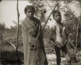 An unidentified young Ho-Chunk boy and girl with smallpox stand outside of their home. The Ho-Chunk girl is wrapped in a shawl and is standing on the left, and the Ho-Chunk boy is posed only in a shirt and is standing on the right.