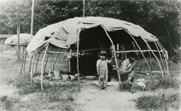 A Winnebago (Ho-Chunk) man and child inside a wigwam with the sides rolled up.