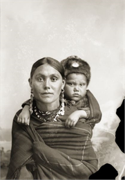 A woman in Ho-Chunk regalia wrapped in a blanket, with a child on her back. The child wears a fur hat, and she has on long earrings, and a necklace. They are posed sitting in front of a painted backdrop.