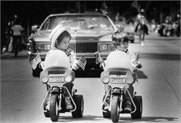 Two toddlers on bigwheels lead memorial day parade. A several cars from the parade are visible behind them.