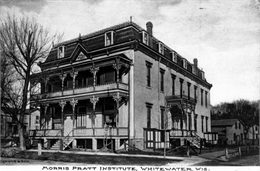 A view of the facade and side of the Morris Pratt Institute. The roof line has an ornate iron railing running around its perimeter and the railings on the porch and balcony are elaborately decorated.