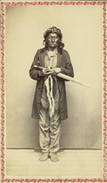 Carte-de-visite studio portrait of a chief standing in front of a backdrop. He is wearing a long coat, fringed shirt, and a crown on his head. In his hands he carries a long-stemmed pipe and a skunk tobacco bag.