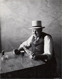 A man sits at a table, holding a cigarette in one hand and pouring a bottle of Tivoli pilsner beer into a glass with the other. Original caption reads: "Special photos taken for Commercial Car Journal to illustrate article on Tivoli Brewery."
