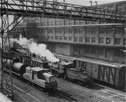 Black and white photo showing an elevated view of Milwaukee railroad yards showing several trains on the tracks next to a large industrial building.