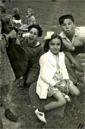 There is a girl sitting on a rock with two boys behind her who are clowning for the camera, and other children around them on the grass.