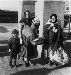 Two immigrant women in headscarves pose with a baby and a young boy. They are both holding baskets and bundles, and are standing on a platform.