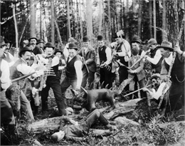 Black and white photograph depicting a staged scene depicting men poised with weapons pointed at a hodag.