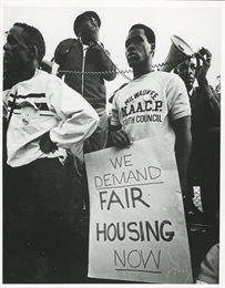 A man holds a sign that reads, "We Demand Fair Housing Now," while others behind him use a bullhorn.