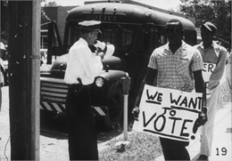 Two men holding protest signs walk down a sidewalk past a white officer who is holding a bullhorn