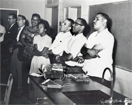 A group of people sing and hold hands while standing at the front of a classroom