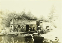 View across water towards the Gang standing in front of G. Swenson's Store in Kettle Falls, which is partially on the water.