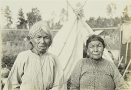 Two elderly women posing in front of a tipi (teepee) at a settlement near Moose River.