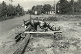 Hodag standing on a board balanced on logs near a road. There is a gasoline filling station in the background.
