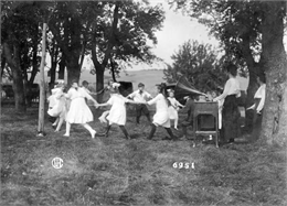 School children dancing in a circle to music from a Victrola phonograph as women look on. The children are outdoors, possibly in a rural schoolyard.