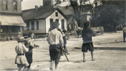 A group of boys "following their leader in search of adventure" in the street. The boys are barefoot and are wearing hats. In the background are a storefront, houses, and wooden wagons.