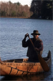 Photograph of Leon C. Valliere, "Ozaawaagosh," wearing a hat and coat and paddling a canoe on a lake
