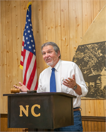 Photograph of Louis V. Clark wearing a white shirt and blue tie, speaking behind a podium and in front of a blue flag