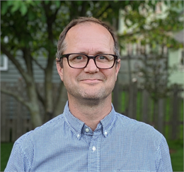 Photograph of Jason Tish, a white man wearing brown glasses and a blue and white checked shirt