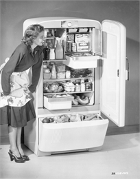 A woman in a long sleeved dress with a floral waist apron leans slightly over to look into a well stocked fridge, with below freezer.