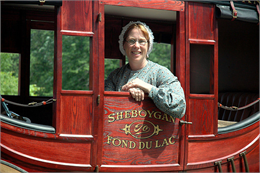 A woman in historical garb sits inside a stagecoach.