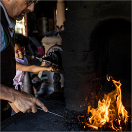 An older man practicing blacksmithing with a younger girl watching