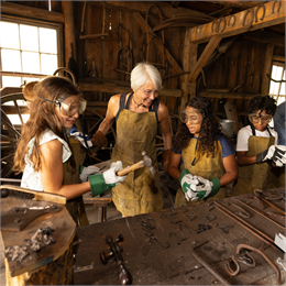 Three younger children and an older woman, all wearing protective gear surrounding a table with metal blacksmithing pieces on it.