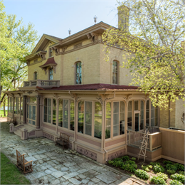 Victorian mansion with ornate trim, large windows, and a red roof sits among trees and gardens, with stone paths and wooden chairs
