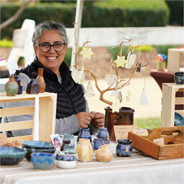 Smiling vendor at an outdoor arts and crafts fair displaying handmade pottery and ornaments on a table.