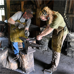 Two people, both wearing protective gear, actively blacksmithing