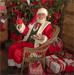 A white man dressed as Santa, holding a cup and a plate of cookies while sitting down