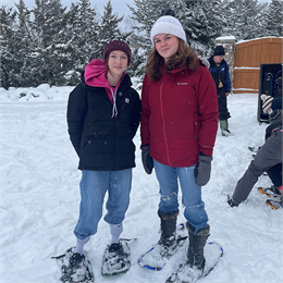 Two young women posing and smiling for the camera. The woman on the left is wearing a dark winter hat and jacket and the women on the right is wearing a white winter hat with a red winter jacket.